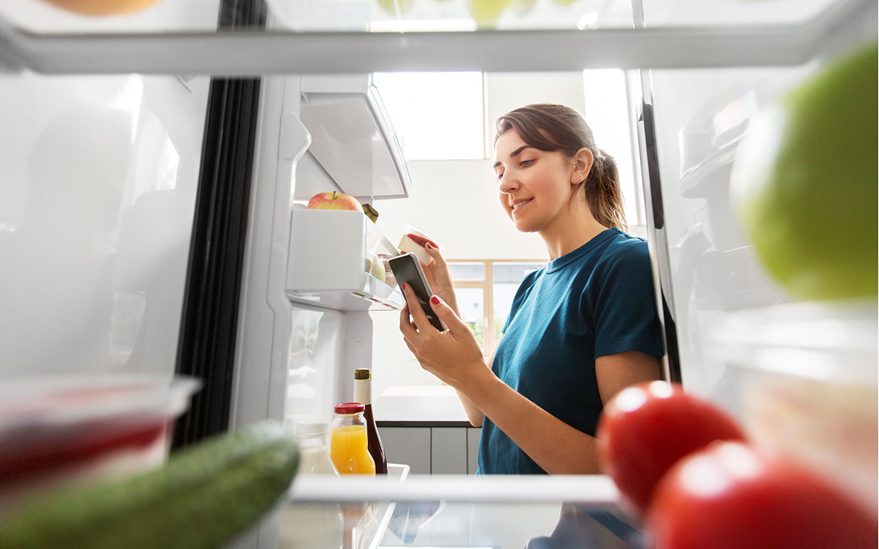 A woman checking her cellphone while taking food from the fridge.